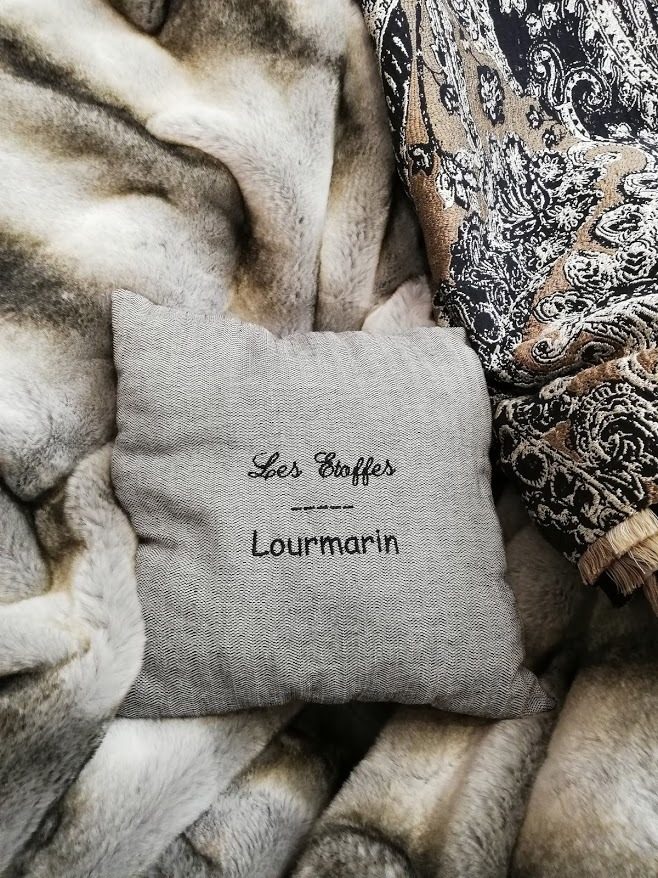 Coussin en lin brodé « Les Etoffes Lourmarin » posé sur un plaid en imitation fourrure