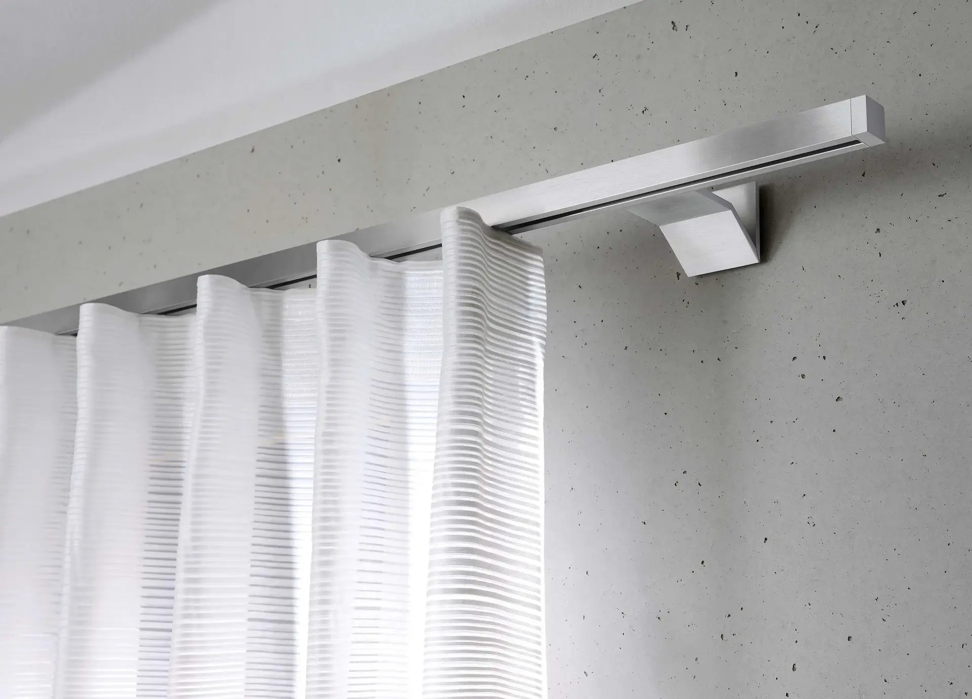 Rail à rideaux en aluminium fixé au plafond portant un voilage blanc à plis réguliers devant un mur en béton